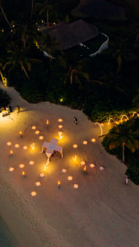 Aerial view of a nighttime beach scene with glowing lanterns arranged in clusters on the sand. Palm trees and dense greenery border the beach, and a small structure with a roof is nestled among the trees. Soft light illuminates parts of the trees and sandy shore, creating a Maldives exclusive experience.