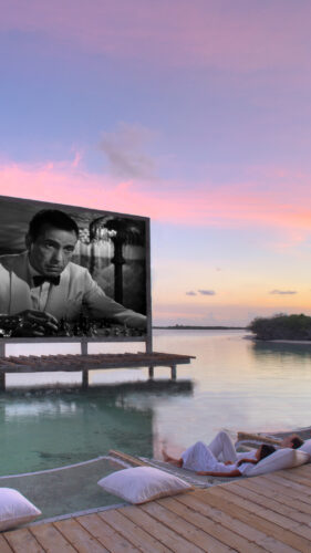 Outdoor movie screening on a floating platform in a serene, tropical setting. The black-and-white film is displayed over calm water, surrounded by a wooden dock with cushions for seating. As the sun sets, painting the sky in soft pinks and purples, indulge in this Maldives exclusive experience at Soneva.