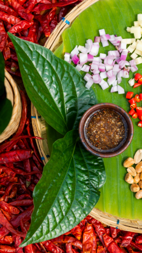 A vibrant food arrangement featuring a small bowl of brown sauce, diced red onions, minced garlic, chopped red chilies, and peanuts on a green banana leaf. As part of Soneva's Thailand Exclusive Experience, two large green leaves lie next to the bowl with a background of dried red chili peppers.