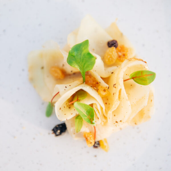 A minimalist gourmet dish at Flying Sauces dining features thinly sliced white vegetables, garnished with microgreens and bits of dried fruit, all artfully arranged on a white speckled plate.