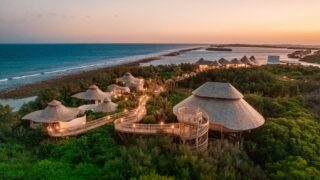 An aerial view of the luxurious, eco-friendly Soneva resort nestled among lush greenery and overlooking a pristine beach at sunset. This Maldives exclusive experience features modern, thatched-roof buildings connected by elevated walkways. The sky is a gradient of warm colors.