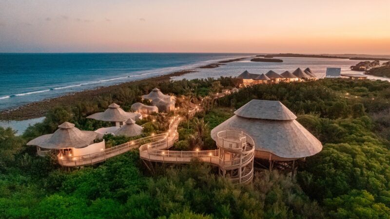 An aerial view of the luxurious, eco-friendly Soneva resort nestled among lush greenery and overlooking a pristine beach at sunset. This Maldives exclusive experience features modern, thatched-roof buildings connected by elevated walkways. The sky is a gradient of warm colors.