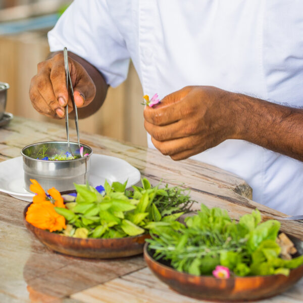 A chef meticulously arranges fresh, garden-picked edible flowers and herbs on a dish using tweezers. Two wooden bowls brimming with vibrant blooms and greens sit beside him. Clad in a white uniform, he stands near a metal tray, perfecting his culinary creation.