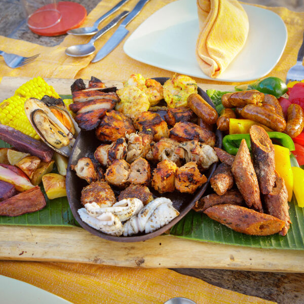 A colorful platter with various grilled meats, sausages, fried plantains, and vegetables such as corn and bell peppers, arranged on a wooden board with a banana leaf. Plates, cutlery, and a napkin are in the background.