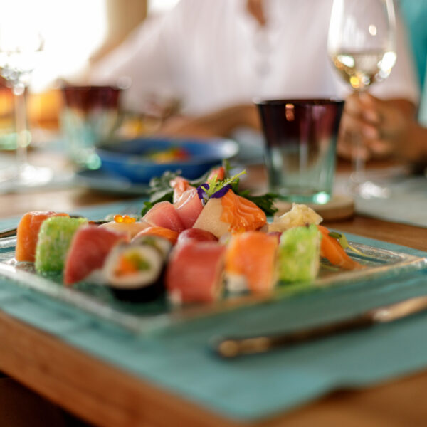 A close-up of a table setting features a glass plate with assorted sushi rolls and sashimi. The scene is accented by Out of the Blue teal placemats, cutlery, and glasses filled with white wine. Blurred figures in the background hint at an elegant dining atmosphere.