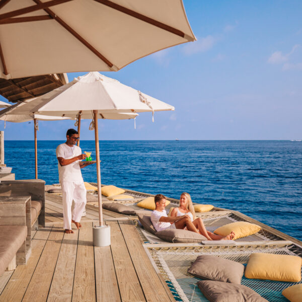 A couple relaxes on lounge nets over the ocean under white umbrellas, while a server in white brings drinks at a luxurious overwater deck with seating and cushions, with clear blue sky and calm sea in the background.