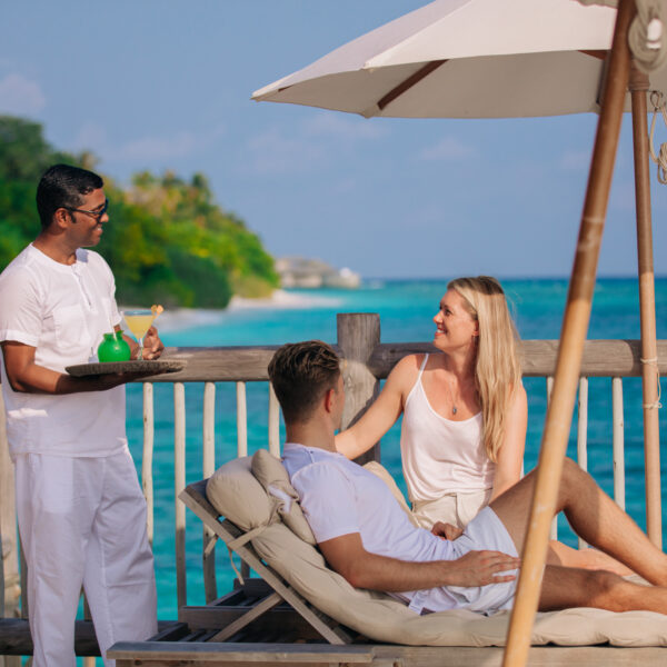 A man and woman relax on lounge chairs under an umbrella by the ocean, while a server in white attire brings them colorful drinks on a tray. The background shows clear blue water and a tropical coastline.