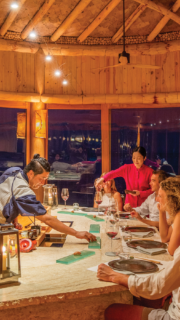 A chef in traditional attire prepares food at a circular sushi bar in a warmly lit, rustic, wooden restaurant. Three patrons sit around the bar with wine glasses, while a server pours wine for a guest. The atmosphere is as cozy and inviting as Maldives dining at Soneva Fushi.