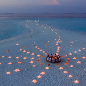 Aerial view of a romantic beach setting at dusk, featuring a winding path of glowing lanterns leading to a central dining area. Encircled by gently lit tents on white sand, the tranquil ocean sets the backdrop for this idyllic escape, showcasing Soneva Exclusive Offers.