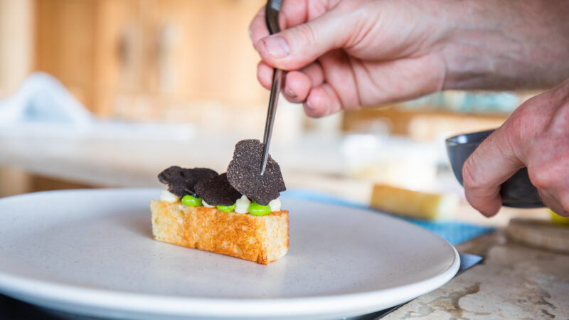A chef uses tweezers to carefully place slices of black truffle on a piece of toasted bread garnished with green peas and cream, all arranged on a plain white plate. The background, reminiscent of a Maldives luxury resort's private villas kitchen, shows a blurred setting.