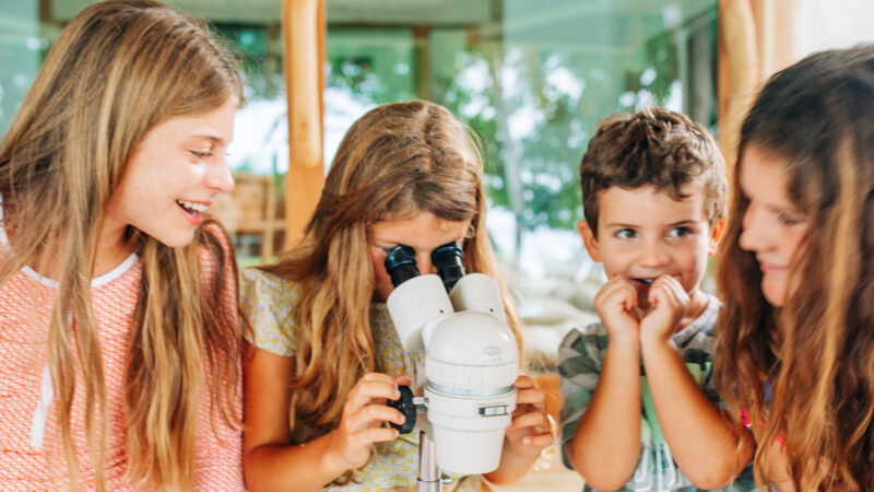 Four children are gathered around a microscope. One girl looks through the eyepiece while the others watch intently. They appear curious and engaged in a bright, indoor setting.