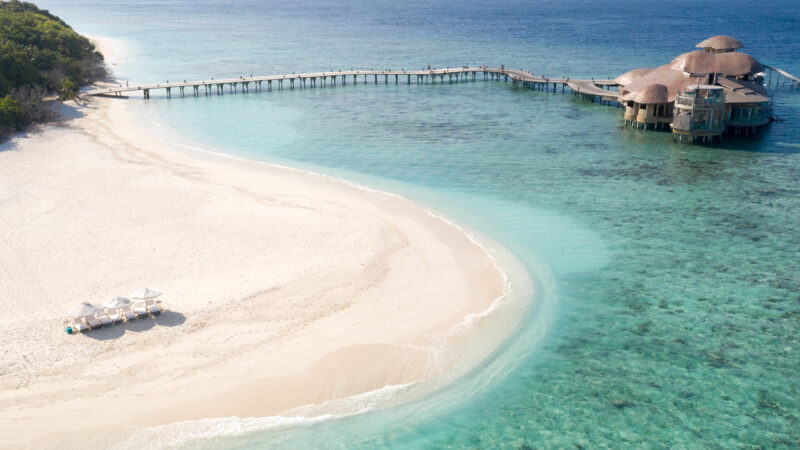 Aerial view of a tropical beach with white sand and clear turquoise water. A wooden pier leads to an overwater structure with thatched roofs. On the beach, two white umbrellas and lounge chairs are set up near the shoreline. Dense greenery borders the sand.