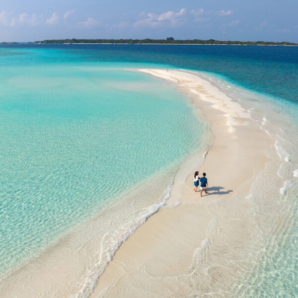 Aerial view of a pristine sandbar with turquoise waters on both sides. A couple walks hand in hand along the sandy path, leading towards a distant lush island against a clear blue sky.