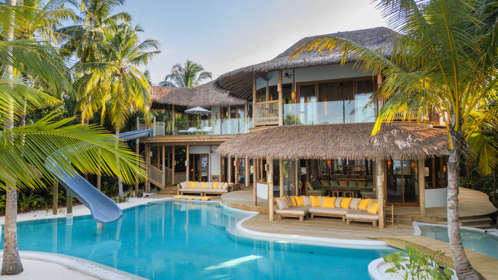 Villa 31 at Soneva Fushi Maldives is a luxurious tropical retreat featuring a thatched roof and glass balconies overlooking a spacious pool. Palm trees surround the area, where a water slide flows into the pool, and yellow cushioned seating lines the serene poolside deck.