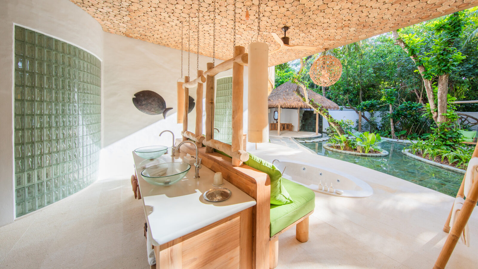Experience the serene luxury of Soneva Fushi Maldives Villa 31's outdoor bathroom, adorned with natural decor. Features include a wooden sink counter, glass bowl sinks, a round mirror, and hanging lights. A freestanding tub overlooks a lush garden and small pool with a thatched-roof backdrop.