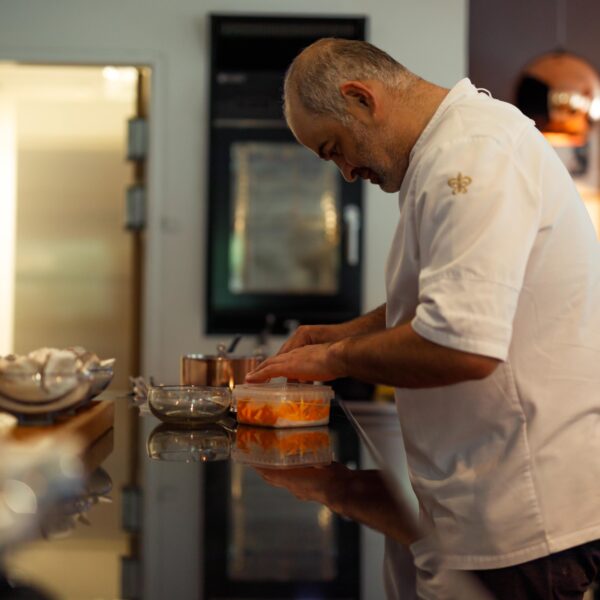 Chef in a white uniform focuses on preparing food in a modern kitchen. He stands at a counter with sliced ingredients, surrounded by culinary tools, with a warm light overhead and framed pictures on the wall.