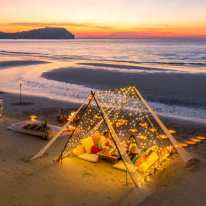 A beach at sunset with a tent adorned with string lights creates the perfect setting to celebrate Eid in the Maldives with Soneva. People relax on cushions inside, surrounded by torches as the sky becomes a gradient of orange and purple, mirrored by the tranquil sea against silhouetted hills.