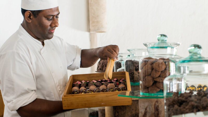 A chef in a white uniform and hat gracefully arranges chocolates in a wooden box at a confectionery shop. Beside him, glass jars brim with assorted chocolates, evoking the sweet allure of spring at Soneva.