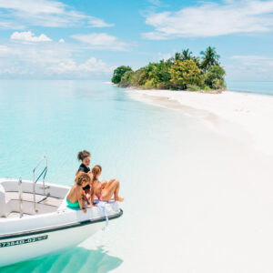 A small, lush island with white sandy beaches and turquoise water is in the background. A motorboat with four people, including two children, is anchored near the shore. The sky is clear with a few clouds.