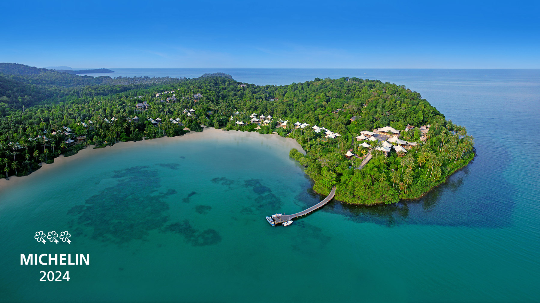 Aerial view of a lush island resort surrounded by turquoise waters. Numerous villas are scattered among dense greenery, with a sandy beach and a curved pier leading from the shore. The image has "Michelin 2024" with an icon in the corner.