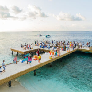 A vibrant crowd gathers on a wooden pier over clear blue ocean waters under a cloudy sky. People are wearing colorful clothing, some holding flags or banners. Two boats are visible in the background.