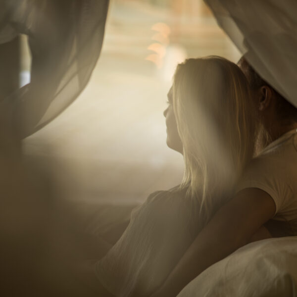 A couple sits closely together on a bed, embraced, and facing a sunlit window. The scene is softly blurred, creating a dreamy and intimate atmosphere. The focus is on their companionship and warmth.