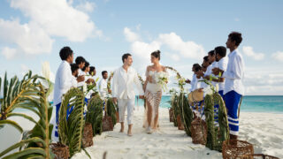 A couple walks hand in hand down a sandy beach aisle lined with tropical leaves and lanterns. They are surrounded by people in white shirts and blue shorts, some holding instruments. The sky is clear with fluffy clouds.