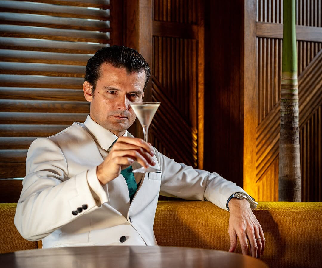 Man in a white suit holds a martini glass while sitting on a yellow sofa. He gazes thoughtfully at the drink. The setting has wooden paneling and subdued lighting, creating an elegant ambiance.