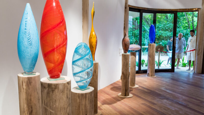 Art gallery featuring Responsibility at Soneva's vibrant glass sculptures on wooden pedestals. Pieces boast blue, red, orange, yellow, and multicolored hues. Large windows frame lush greenery as two people admire from outside. Gleaming wooden floors and posts complete the scene.