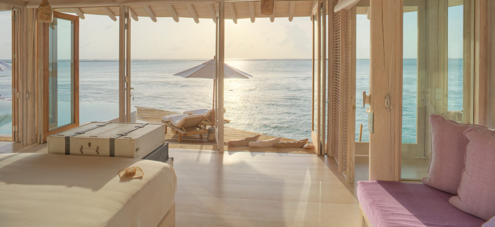 A serene beachfront villa with open sliding glass doors revealing an infinity pool and ocean view. Sunlight reflects on the water, while two lounge chairs with a beach umbrella sit on a wooden deck. Soft pastel decor enhances the tranquil atmosphere.