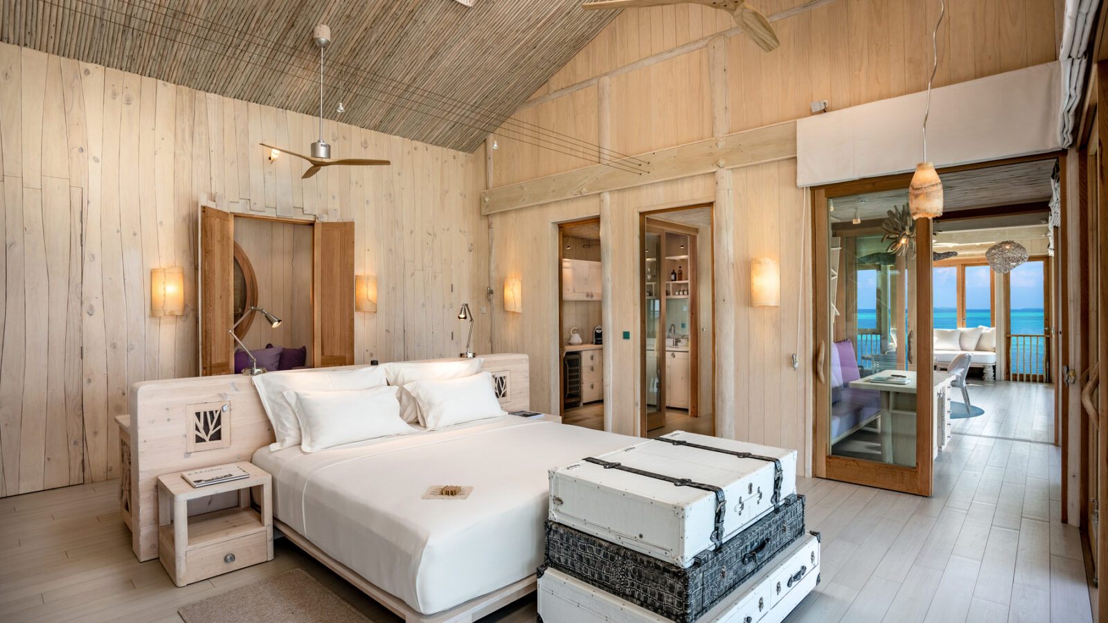Luxurious beach resort room with a king-size bed, white linens, and wooden decor. Large windows reveal a view of the ocean. The room includes ceiling fans, pendant lights, and wicker furniture, creating a serene and tropical ambiance.