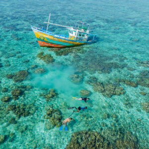 Aerial view of a vibrant, colorful boat anchored in clear turquoise water at Soneva Secret, surrounded by coral reefs. Four snorkelers explore the shallow waters below, revealing the vivid marine life and structures—a signature experience that captivates every adventurer.