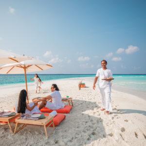 Three people relax on cushioned loungers under umbrellas on a sandy beach by turquoise water during summer at Soneva Fushi, while a server in white carries a tray of food toward them. Another person in white strolls along the shore in the background.