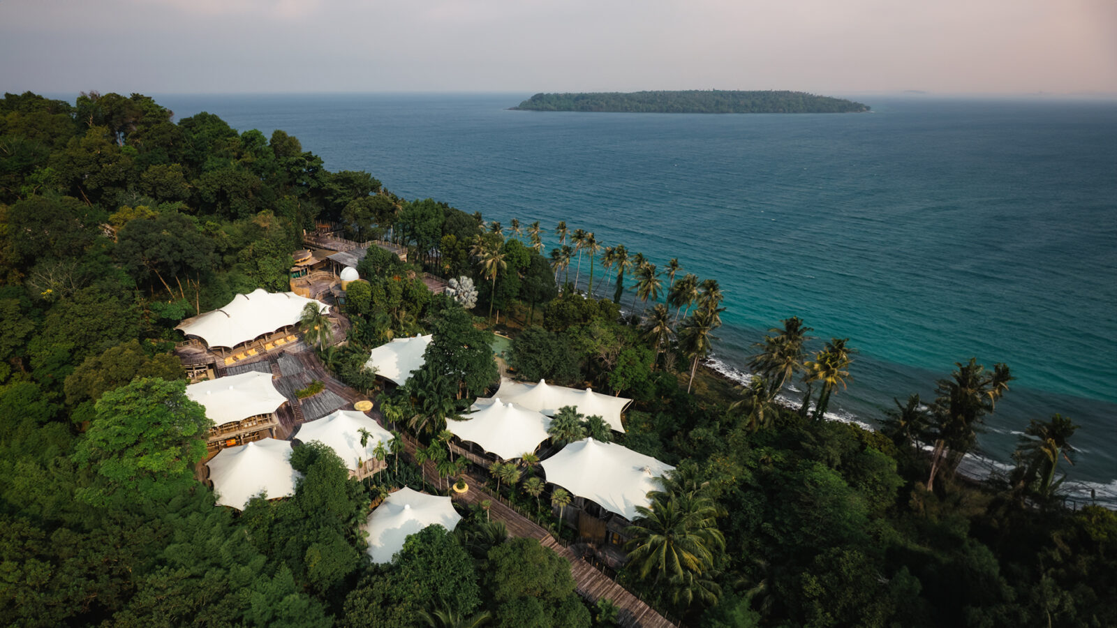 Aerial view of a tropical coastal resort with white tented structures nestled among lush green trees beside a blue ocean—capturing the essence of summer at Soneva Fushi, with a distant island visible on the horizon under a hazy sky.