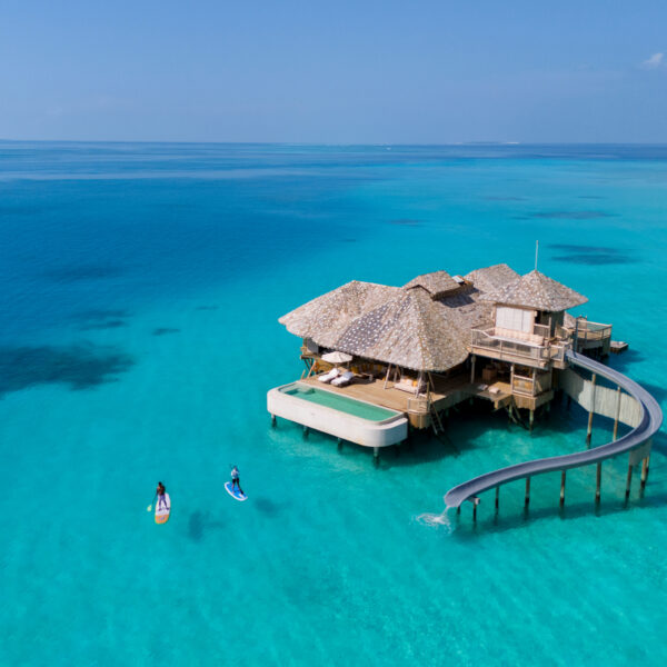 A thatched-roof overwater villa with a waterslide sits above clear turquoise ocean. Two people paddleboard nearby under a bright blue sky.