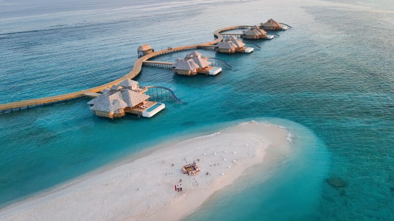 Aerial view of overwater villas connected by a wooden walkway above turquoise ocean waters, with a small sandy island and a group of people gathered on the beach below.
