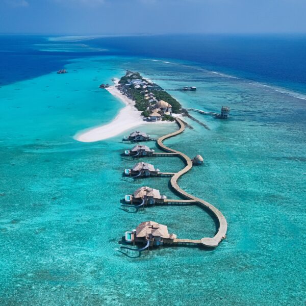 Aerial view of overwater villas connected by a winding wooden walkway leading to a small island surrounded by turquoise ocean and coral reefs. The island has white sandy beaches and lush greenery.