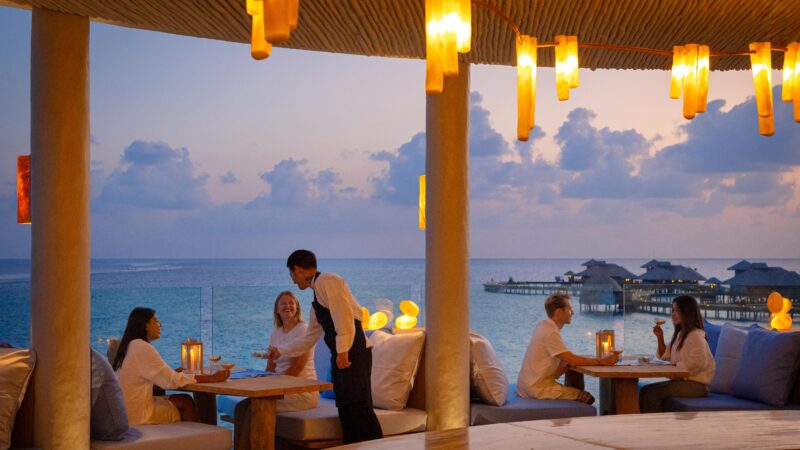 People dine at an outdoor restaurant overlooking the ocean at sunset. Warm hanging lights create a cozy atmosphere, while a waiter serves guests. Overwater bungalows are visible in the background.