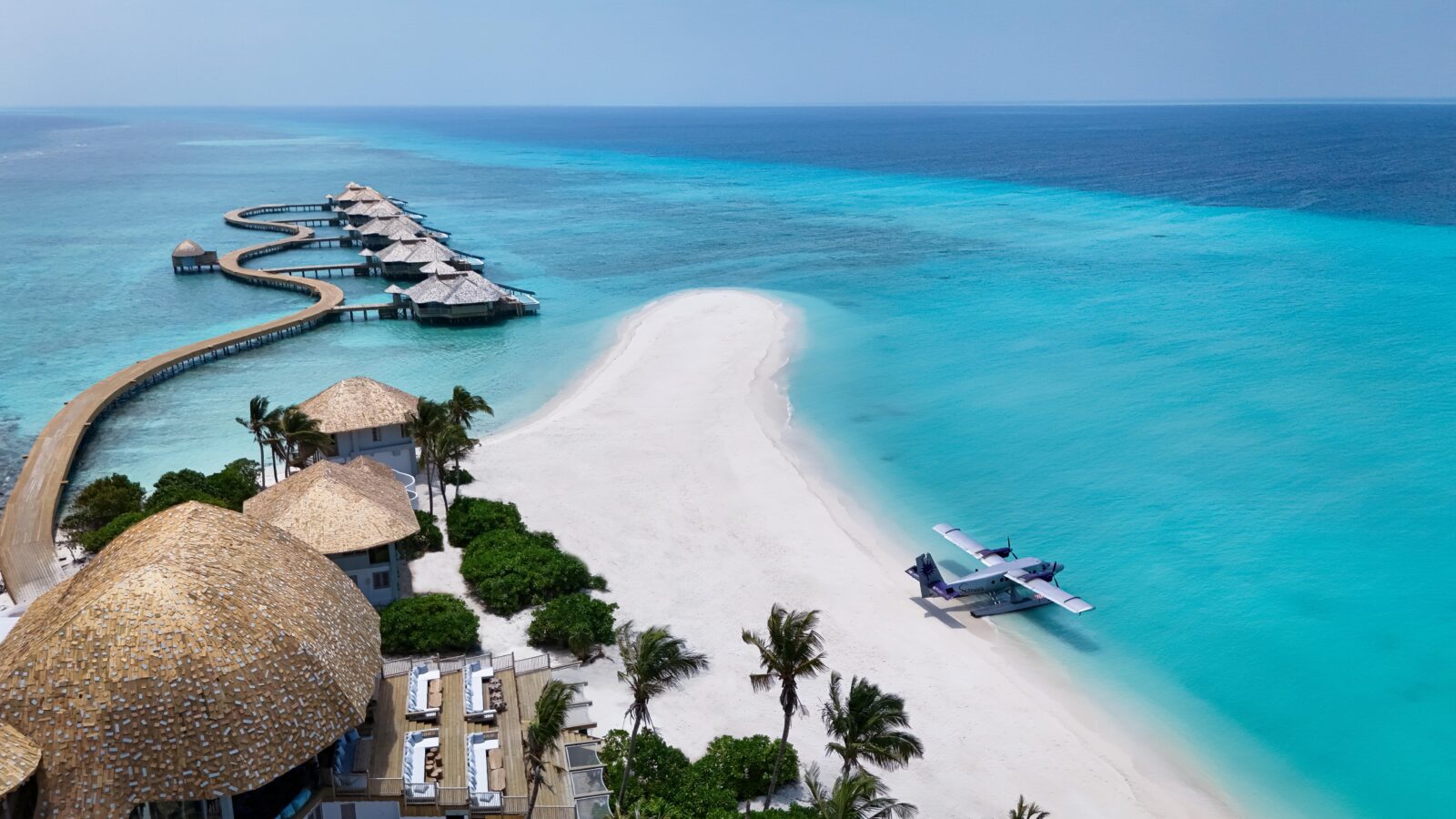Aerial view of a tropical island with overwater bungalows, a sandy beach, turquoise ocean, lush palm trees, and a small seaplane parked near the shore.