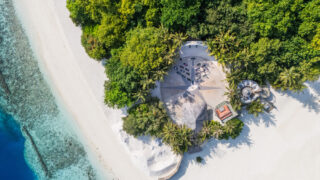 Aerial view of a tropical beach with white sand, turquoise water, dense green trees, and the Soul at Soneva resort area featuring shaded structures and lounge chairs nestled among swaying palm trees.