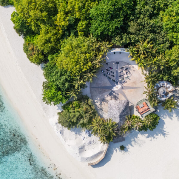 Aerial view of a tropical beach with white sand, turquoise water, dense green trees, and the Soul at Soneva resort area featuring shaded structures and lounge chairs nestled among swaying palm trees.