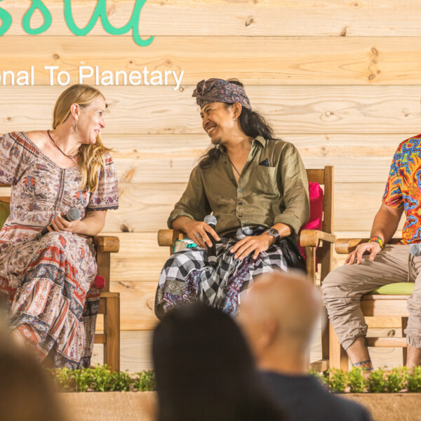 Three people sit smiling and talking on a stage with a wooden background at Soul at Soneva. The woman and man in the center are engaged in conversation, while the man on the right looks forward, smiling. Audience members are visible in the foreground.