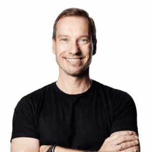 A smiling man with short light brown hair and a trimmed beard, wearing a black t-shirt and bracelets, stands with arms crossed against a plain white background, embodying the relaxed spirit of Soul at Soneva.