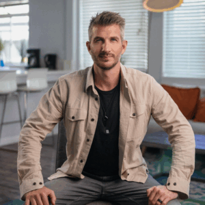 A man with short, spiked hair and a trimmed beard sits indoors on a chair at Soul at Soneva. He wears a beige button-up shirt over a black t-shirt and gray jeans, set against a bright kitchen and inviting living area.
