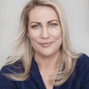 A woman with long blonde hair and blue eyes, wearing a navy blue top and gold necklace, smiles gently at the camera against a light, neutral background—radiating tranquility reminiscent of Soul at Soneva.