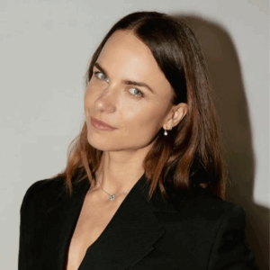 A woman with straight brown hair, light eyes, and fair skin wears a black blazer and a small pendant necklace, standing against a light background at the Soul at Soneva event and smiling slightly at the camera.