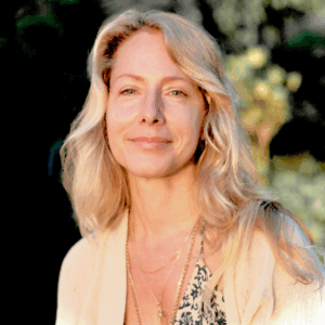 A woman with long, wavy blonde hair smiles softly outdoors at Soul at Soneva. She wears a light-colored cardigan and layered necklaces, with sunlight illuminating her face and a blurred green background behind her.