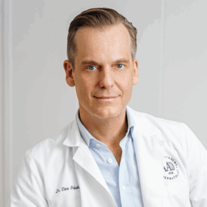 A middle-aged man with light hair wearing a white medical coat and a light blue shirt, smiling gently while facing the camera against a plain background—a professional at Soul at Soneva.