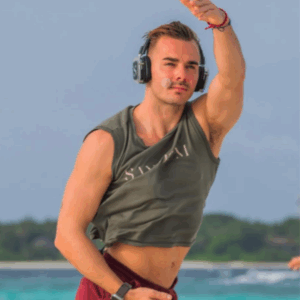 A man wearing headphones and a sleeveless shirt is dancing or exercising outdoors near the sea at Soul at Soneva, lifting one arm and looking focused. He has a microphone headset and wristband, with blue water and sky blurred in the background.