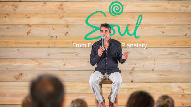 A barefoot man in a dark shirt and light pants sits on a stool, speaking into a microphone. He is in front of a wooden wall with "Soul at Soneva" and the text "From Personal to Planetary." Audience heads are visible.